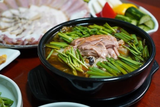검은 냄비에 끓인 붉은빛 국물의 탕 — 둘레에 푸른 채소, 가운데 분홍빛 건더기, 뒤로 홍어·수육 접시 일부