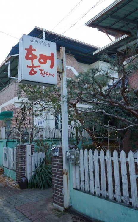 ‘홍어전문점 홍어한마리’ 돌출 간판이 걸린 벽돌 2층 집과 흰 나무 울타리, 소나무